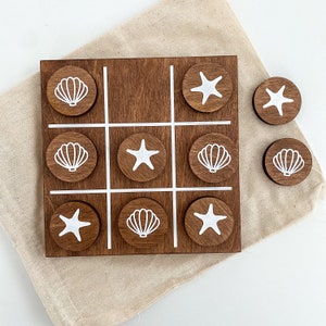 May include: A wooden tic-tac-toe game with a nautical theme. The game board is brown and features images of seashells and starfish. The game comes with nine playing pieces, five of which are shown in the image.