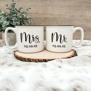 Puede incluir: Dos tazas de cer&aacute;mica blanca con bordes y asas negros. La taza de la izquierda dice "Mrs. 03.29.25" y la taza de la derecha dice "Mr. 03.29.25".