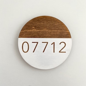 Könnte beinhalten: Ein rundes Holzschild mit einer braunen oberen Hälfte und einer weißen unteren Hälfte. Die Nummer 07712 ist in weiß auf der weißen Hälfte des Schildes gedruckt.