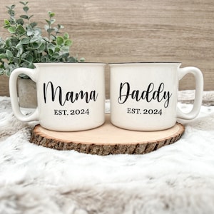 May include: Two white ceramic mugs with black handles and black text. The mug on the left says "Mama EST. 2024" and the mug on the right says "Daddy EST. 2024".