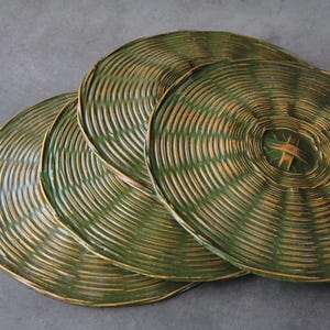 May include: Four round, woven placemats in shades of green and tan are arranged on a gray surface. The placemats have a concentric circular design, with a central woven detail. The natural materials and colors suggest a rustic, eco-friendly theme.