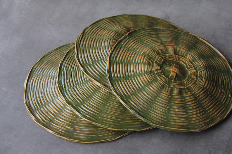 May include: Four round, woven placemats in shades of green and tan are arranged on a gray surface. The placemats have a concentric circular design, with a central woven detail. The natural materials and colors suggest a rustic, eco-friendly theme.