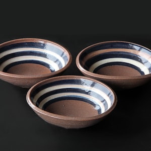 May include: Three brown ceramic bowls with white and blue striped designs. The bowls are arranged in a triangular pattern on a black surface.