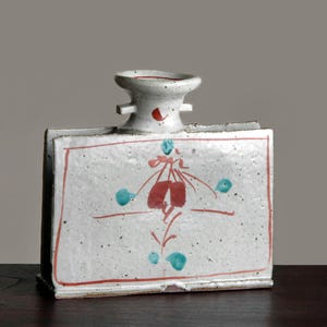 May include: A rectangular ceramic vase with a white base, red trim, and a floral design in red and teal. The vase has a cylindrical top with two small handles. The vase is on a dark wooden surface.