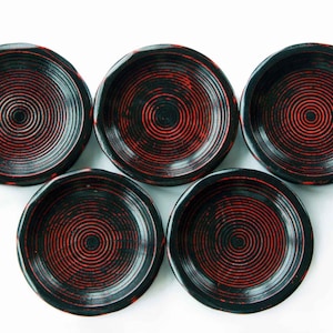 May include: Five small, round, black dishes with a red spiral design in the center. The dishes have a slightly wavy edge and are arranged in a circular pattern against a white background. The design is reminiscent of traditional Japanese lacquerware.