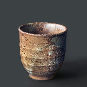 Puede incluir: Una taza de cerámica hecha a mano con un aspecto terroso texturizado. La taza presenta bandas horizontales de color marrón, beige y blanco, con un borde ligeramente ensanchado. La taza mide unos 10 cm de alto.