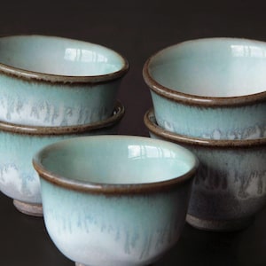 May include: Six ceramic bowls with a pale blue glaze and a brown rim. The bowls are stacked in pairs and arranged on a dark surface. The bowls have a simple, elegant design.