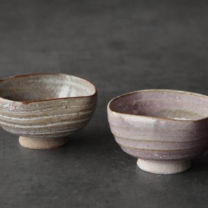 Puede incluir: Dos cuencos de cerámica hechos a mano. Uno es marrón claro con rayas horizontales blancas y beige. El otro es de color púrpura claro con rayas horizontales. Ambos tienen una base pequeña y redonda.