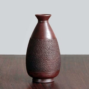 May include: A dark brown vase with a textured, patterned lower half and a smooth, tapered upper portion. The vase sits on a dark wooden surface, with a light gray background.