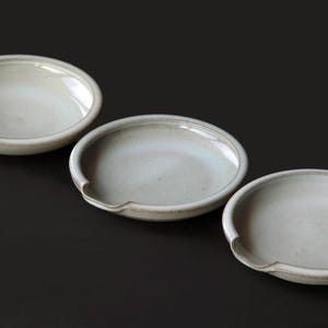 May include: Three off-white ceramic spoon rests with a curved design. The spoon rests are oval-shaped with a small, curved indentation on one side. The ceramic has a glossy finish and a subtle, textured appearance.