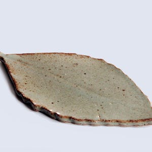 以下が含まれることがあります： 明るい緑色の釉薬と質感のある表面を持つ、葉の形をしたセラミック皿。縁は濃い茶色です。この皿は葉に似せてデザインされています。