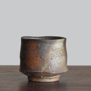 Może przedstawiać: Ręcznie wykonany ceramiczny kubek o unikalnym, asymetrycznym kształcie. Kubek ma ciemnobrązową powierzchnię zewnętrzną z jaśniejszymi, teksturowanymi obszarami i lekko nieregularnym brzegiem. Podstawa jest jaśniejsza, dodając kontrastu.