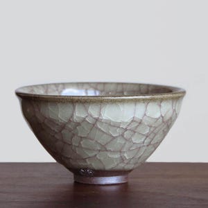 May include: A ceramic bowl featuring a light sage green glaze with a prominent dark brown crackle pattern across the exterior surface. The bowl has a smooth, rounded rim and a dark, unglazed brown base that tapers toward the bottom.