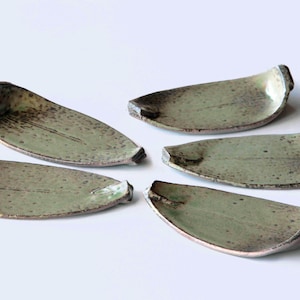 Op de afbeelding: Vijf handgemaakte keramische bladvormige schalen. De schalen hebben een gespikkelde, gedempte groene glazuur met donkerbruine accenten. Elke schaal heeft een verhoogde, gebogen rand aan één uiteinde en ze zijn gerangschikt op een wit oppervlak.