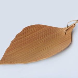 以下が含まれることがあります： 木製の葉の形をしたサービングトレイ。自然な木目仕上げで、明るい茶色です。編まれた素材で作られた小さなハンドルが付いています。トレイの端はわずかに不均一です。