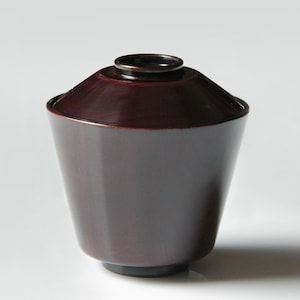 May include: A dark burgundy lidded bowl with a conical shape. The lid is a darker shade of burgundy than the bowl's body. The bowl sits on a small, dark base. The design is simple and elegant, suitable for serving.