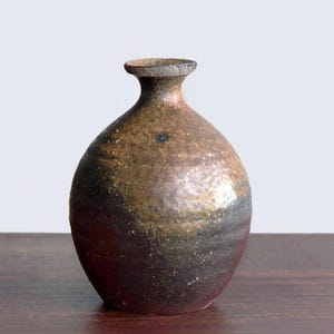 May include: A handcrafted, brown ceramic vase with a rounded body and a narrow neck. The vase has a textured surface with dark brown, tan, and black hues. The vase is displayed on a wooden surface.