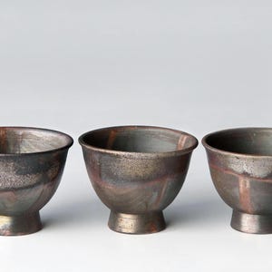 Könnte beinhalten: Drei kleine Keramikschalen mit einer dunklen, metallischen und schillernden Oberfläche. Jede Schale hat einen abgerundeten Körper, eine weite Öffnung und einen kurzen, stabilen Standfuß. Die Oberfläche zeigt gesprenkelte Töne in Anthrazit, Bronze und Kupfer.