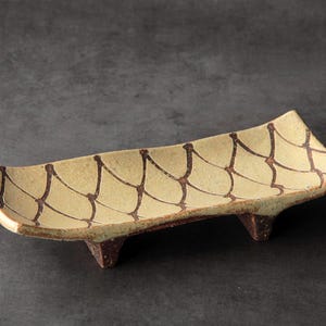 May include: A rectangular ceramic dish with a light yellow base and brown geometric pattern. The dish is raised on three small, dark brown feet. The edges are slightly curved, and the overall design is simple and elegant.