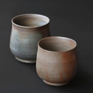 Puede incluir: Dos tazas de cerámica con forma redondeada y abombada y base con pie. La taza en primer plano presenta un esmalte cálido y terroso en tonos terracota y bronceado, mientras que la taza detrás muestra un acabado gris y marrón más frío.