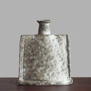 Puede incluir: Una botella de cerámica plana y rectangular con un cuello estrecho y una base ancha y acampanada. La superficie presenta un esmalte texturizado y moteado en tonos blanco roto, gris y marrón oscuro con un acabado rugoso.