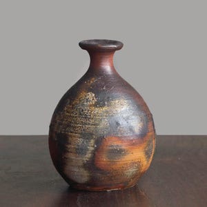 Könnte beinhalten: Eine kleine, runde Keramikvase mit engem Hals und ausgestelltem Rand. Die Oberfläche weist ein gesprenkeltes, strukturiertes Finish mit vermischten Tönen aus Dunkelbraun, Anthrazit und gebranntem Orange sowie horizontalen Rillen auf.