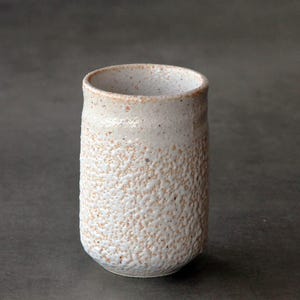 Puede incluir: Un vaso cilíndrico de cerámica con un exterior texturizado y moteado en tonos blanco roto y bronceado claro. La superficie presenta un acabado rugoso y granular que se transforma en un borde más suave y claro en la parte superior.