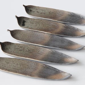 May include: Five silver-toned, leaf-shaped decorative trays arranged diagonally on a white surface. Each tray has a textured surface with a slightly curved shape and a small, raised edge at one end. The trays are empty.