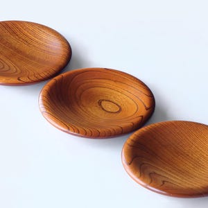 Puede incluir: Tres cuencos redondos de madera, de color marrón cálido y con vetas visibles. Estos cuencos poco profundos y curvados son ideales para servir pequeñas porciones. El acabado de madera natural les da un aspecto rústico y elegante.