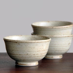 Puede incluir: Un juego de cuatro cuencos de cerámica con un aspecto rústico y artesanal. Los cuencos son de color blanco roto con motas marrones y líneas horizontales. Tienen forma redondeada y están sobre una superficie de madera oscura.
