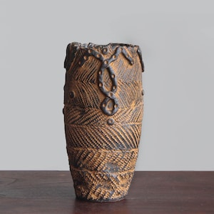 Peut inclure: Un vase cylindrique en céramique présentant une surface texturée avec une palette de couleurs brun foncé et beige. L'extérieur affiche des motifs gravés en diagonale complexes et des détails linéaires en relief brun foncé.