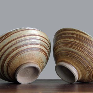 Może przedstawiać: Dwie ceramiczne miski o unikalnym designie. Miski są beżowe z brązowymi poziomymi paskami. Miski są pochylone, ukazując wnętrze i podstawę. Miski znajdują się na drewnianej powierzchni na szarym tle.