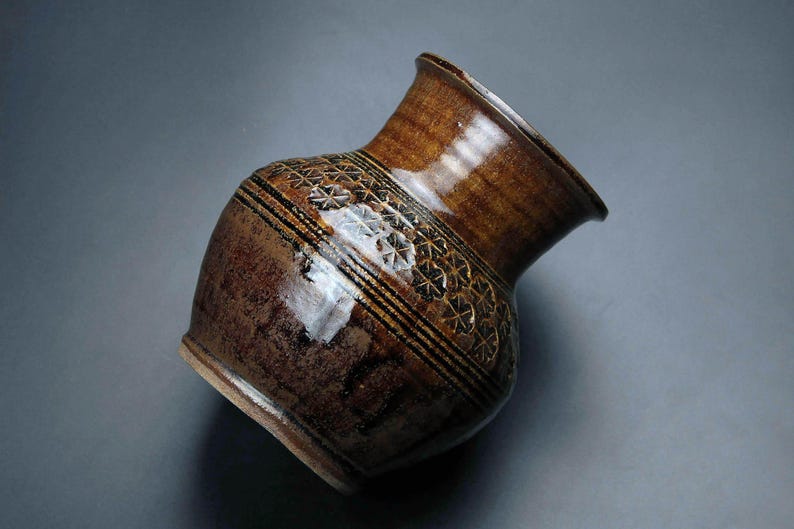 K&ouml;nnte beinhalten: Eine braune Keramikvase mit einem geometrischen Muster in schwarzer Glasur. Die Vase hat eine breite &Ouml;ffnung und einen runden K&ouml;rper.