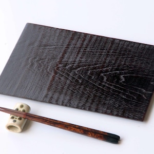 May include: A dark brown wooden serving board with a rectangular shape and visible wood grain. A pair of chopsticks and a ceramic chopstick rest are placed on the white surface next to the board. The chopsticks are brown with black tips.