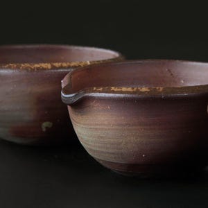 May include: Three handmade, rustic ceramic bowls in varying shades of brown and reddish-brown. Each bowl has a small spout and a textured surface. The bowls are arranged in a row, with the focus on the two closest bowls.