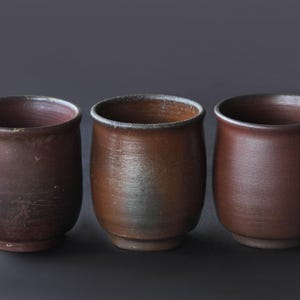 May include: Three cylindrical ceramic cups with a matte, dark brown finish. Each cup features a slightly flared rim, a rounded body, and a small, raised circular base. The surfaces show subtle variations in texture and earthy brown tones.