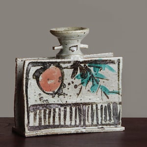 May include: A handcrafted ceramic vase with a unique, rectangular shape. The vase features a light-colored base with black, red, and teal painted designs. The top has a small, round opening and two small handles. The vase is approximately 8 inches tall.