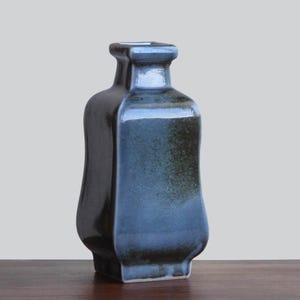 Puede incluir: Un jarrón de cerámica con cuerpo cuadrado y cuello estrecho. El jarrón tiene un esmalte azul oscuro brillante con toques de marrón y negro. El jarrón se exhibe sobre una superficie de madera.