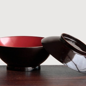 May include: A black and red lacquered bowl with a matching lid. The bowl has a glossy finish, with a red interior and a black exterior. The lid is partially off to the side. The bowl is sitting on a dark wooden surface.