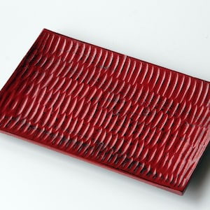 May include: A rectangular, red serving tray with a textured, carved surface. The tray's design features a repeating pattern of curved lines, creating a visual texture. The tray is a deep red colour.