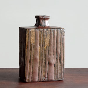 Pode incluir: Um vaso retangular de cerâmica com um design listrado vertical texturizado. O vaso tem um esquema de cores marrom e bege com uma pequena abertura quadrada no topo. O vaso está sobre uma superfície de madeira.