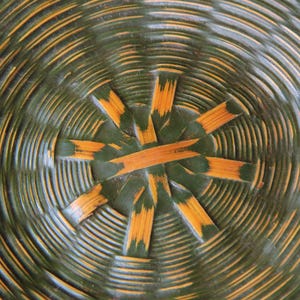 May include: Close-up of a woven basket base. The basket features concentric circles in shades of green and yellow-orange. The center has a starburst design with radiating segments.