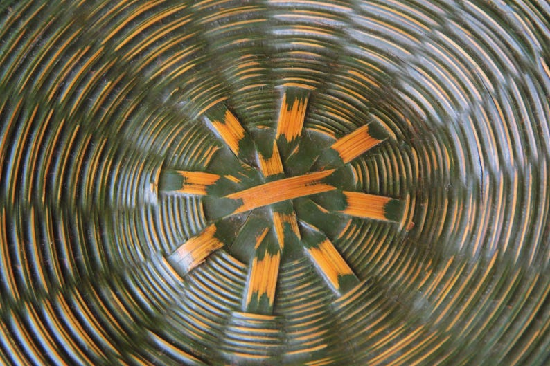 May include: Close-up of a woven basket base. The basket features concentric circles in shades of green and yellow-orange. The center has a starburst design with radiating segments.