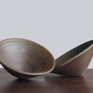 May include: Two small, conical bowls with a dark, textured, matte brown finish. The bowls have a simple, unadorned design with a smooth interior surface and a slightly rougher exterior texture, showcasing a deep, earthy tone throughout the pieces.