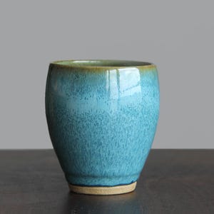 May include: A ceramic cup with a rounded body and a tapered base, featuring a glossy glaze in shades of turquoise and light blue. The rim and the base are finished in a contrasting earthy brown tone, showcasing a speckled texture throughout.
