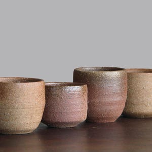 May include: A set of four small, cylindrical ceramic cups with a rough, textured surface. The cups feature earthy, mottled tones of brown, tan, and reddish-clay hues, each with a slightly different height and color variation in the finish.
