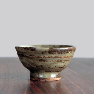 May include: A small, handcrafted ceramic bowl with a footed base. The bowl has a speckled, earthy green glaze with horizontal bands of darker brown. The interior is a lighter green. The bowl sits on a dark wooden surface.