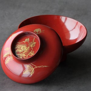 Könnte beinhalten: Eine rot lackierte Schüssel mit passendem Deckel. Der Deckel zeigt ein goldenes Blumenmuster. Die Schüssel und der Deckel haben eine glänzende Oberfläche. Die Schüssel ist offen und gibt ihr Inneres frei. Das Design ist wahrscheinlich japanischen Ursprungs.