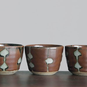 Puede incluir: Tres tazas de cerámica hechas a mano con un esmalte marrón y patrones abstractos blancos y verde claro. Las tazas son cilíndricas con un borde ligeramente ensanchado y se asientan sobre una base de color beige claro. Las tazas se exhiben sobre una superficie de madera oscura.