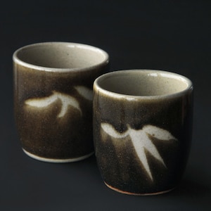 Peut inclure: Deux tasses en céramique avec un glaçage brun foncé et des motifs blancs en forme de feuilles. Les tasses ont une forme cylindrique avec un bord légèrement évasé et un intérieur de couleur claire. Les tasses sont probablement destinées à boire du thé ou d'autres boissons.